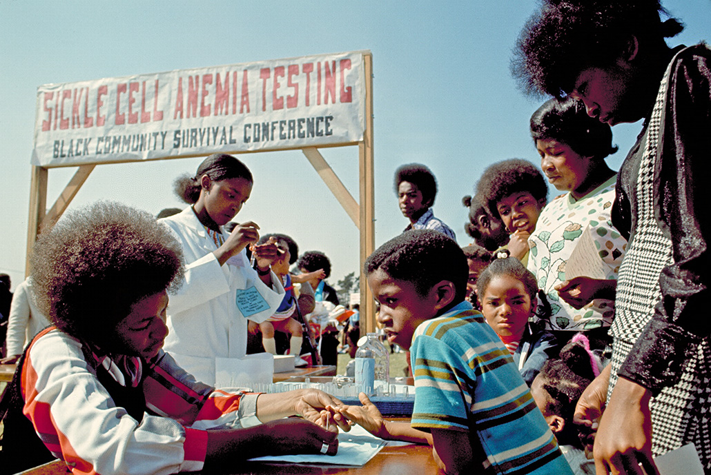 A vintage photograph of a Sickle Cell Anemia Testing site hosted by the Black Panther Party. Children are getting tested while standing close to their parents. There's a banner behind the testers that reads: "SICKLE CELL ANEMIA TESTING Black community survival conference"